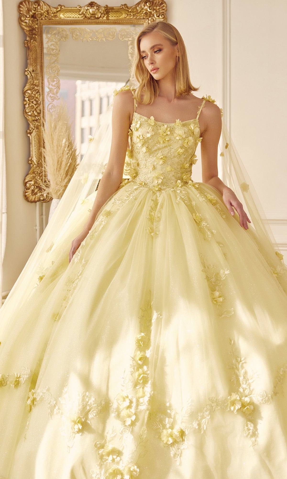 A woman wearing the Juliet 1436 Glitter Quinceañera Dress with Cape stands indoors near a large ornate gold mirror.